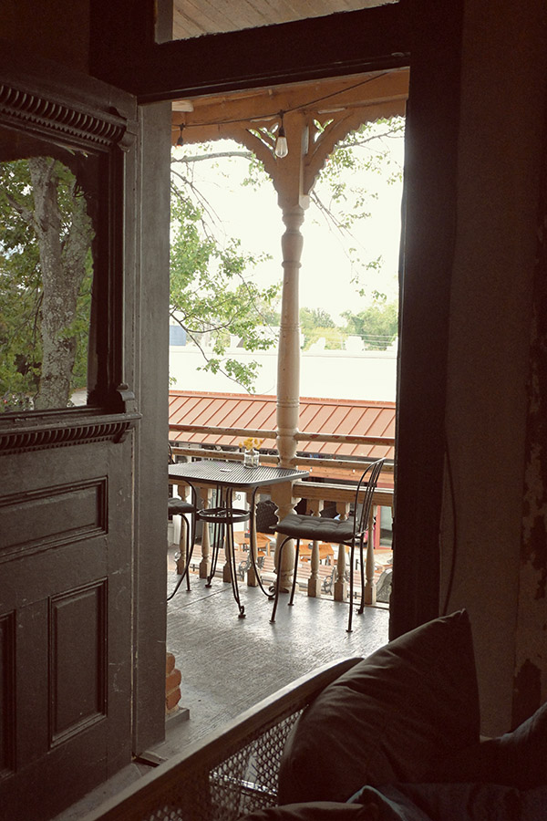 French-country-design-style-of-a-covered-balcony-with-a-black-metal-bistro-table-and-two-chairs-a-light-colored-wooden-baluster