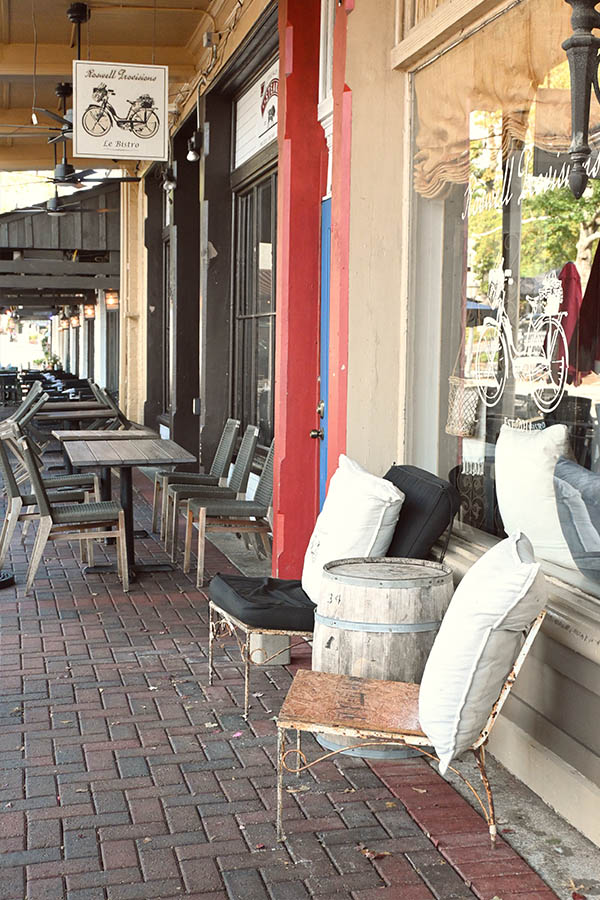 Roswell-Provisions-with-a-red-door-and-windows-and-outdoor-seating-on-a-brick-pavement-in-front-of-it-including-two-chairs-and-a-small-wooden-barrel