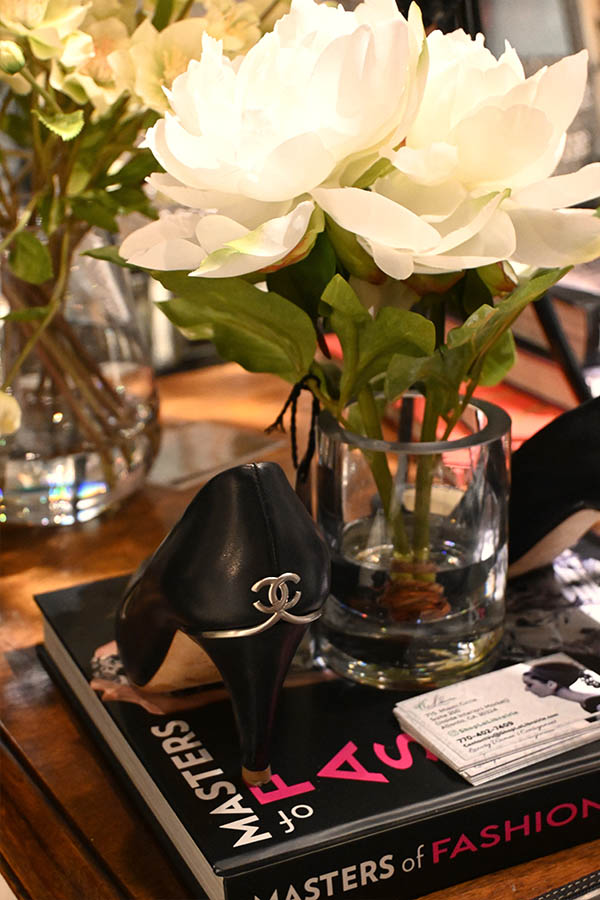 black-high-heeled-shoe-with-a-gold-Chanel-CC-logo-on-the-heel-is-resting-on-a-book-titled-“MASTERS-of-FASHION”-next-to-a-glass-vase-of-white-flowers