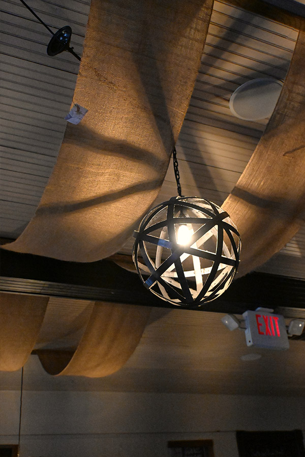 ceiling-with-a-decorative-metal-orb-shaped-light-fixture-that-has-an-exposed-bulb-and-it-is-hanging-from-a-chain-in-the-center-of-the-frame-with-two-swags-of-burlap-like-fabric