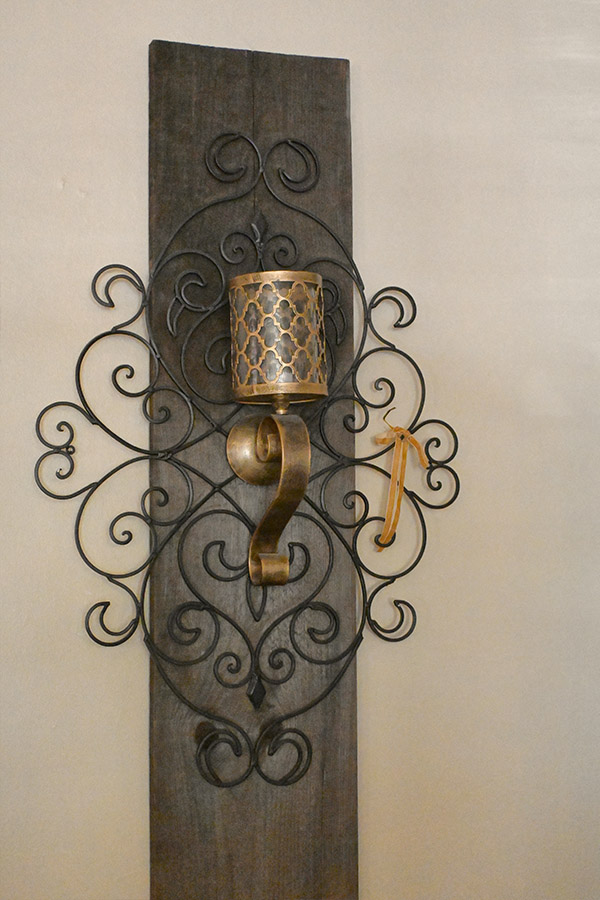 dark-wooden-plank-backing-and-ornate-black-metal-scrollwork-that-features-a-gold-colored-candle-holder-with-a-geometric-patterned-shade
