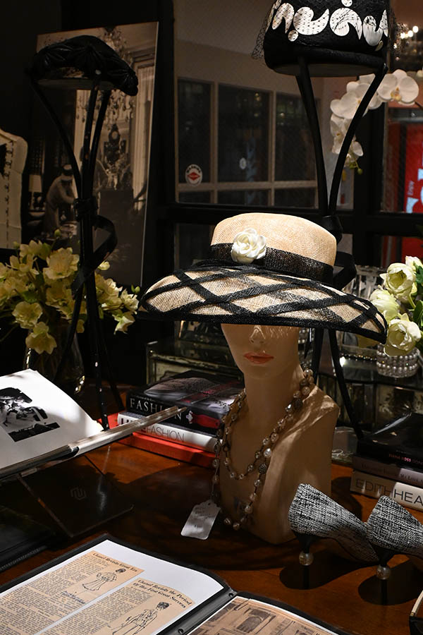 display-is-shown-with-a-mannequin-head-wearing-a-wide-brimmed-straw-hat-with-a-black-ribbon-and-white-flower,-surrounded-by-books-and-other-accessories
