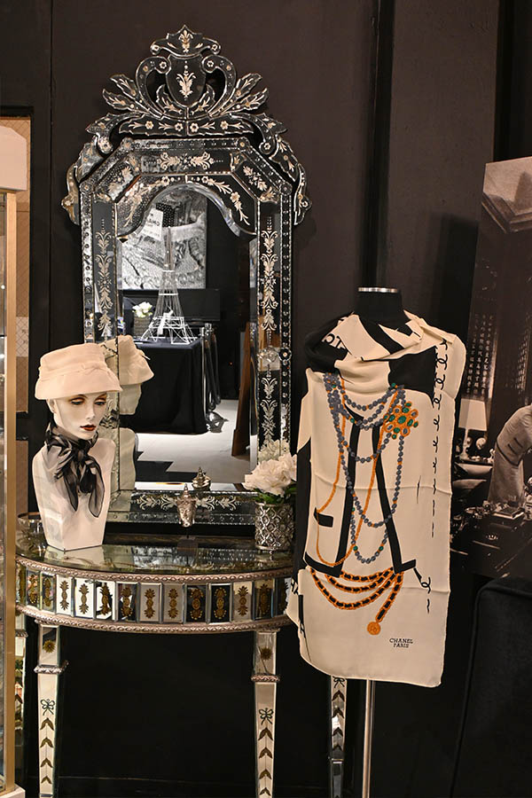 display-is-shown-with-an-ornate-mirrored-dressing-table,-a-head-mannequin-wearing-a-hat-and-scarf,-and-a-torso-mannequin-wearing-a-Chanel-scarf-that-features-an-illustration-of-a-jacket-and-jewelry