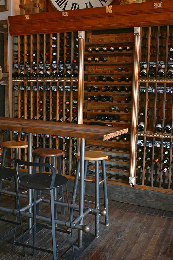 french-cafe-wooden-wine-rack-display-with-many-bottles-of-wine-and-a-long-wooden-bar-table-in-front-of-it-with-several-metal-stools-and-the-wine-racks