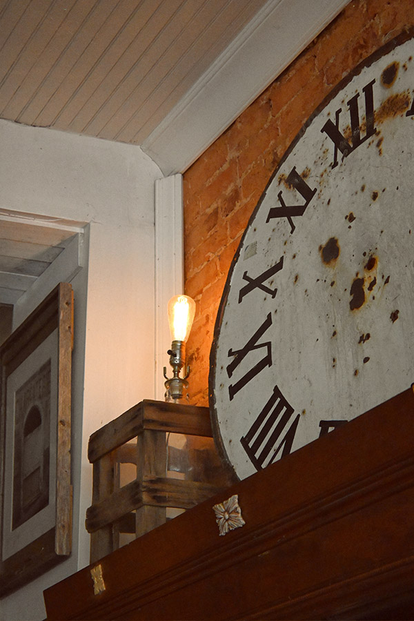 french-interior-space-with-a-large-weathered-white-clock-face-with-black-Roman-numerals-leaning-against-a-brick-wall-next-to-a-small-wooden-crate-holding-a-single-lightbulb-on-a-lamp