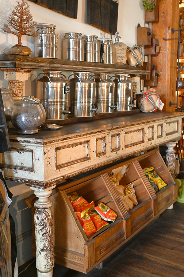 french-rustic-and-distressed-white-wooden-counter-or-display-unit-with-multiple-silver-canisters-of-varying-sizes-on-the-shelves