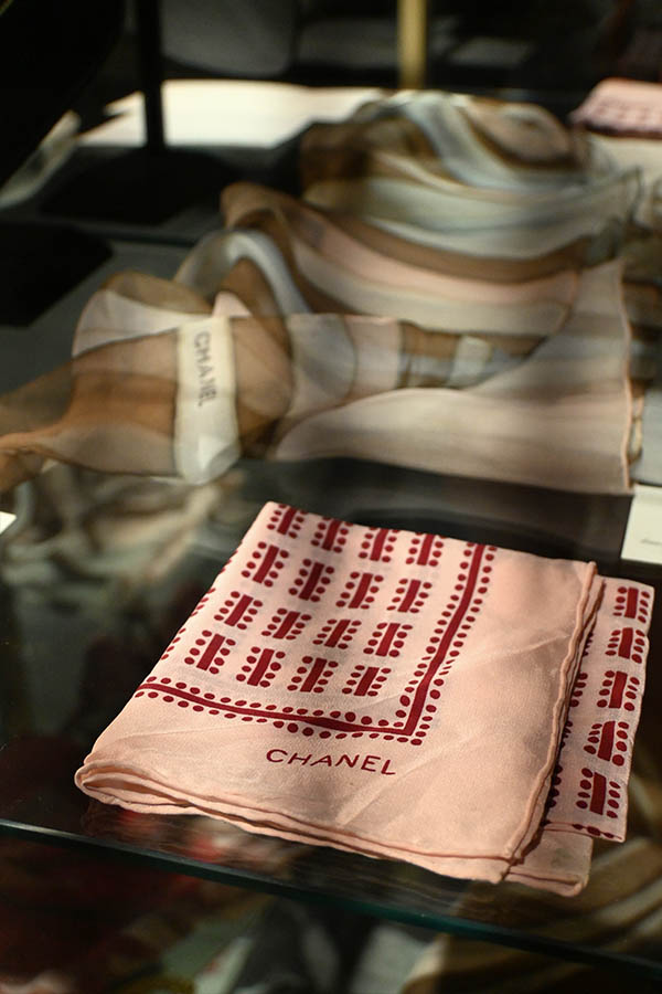 glass-display-case-is-shown-with-two-Chanel-scarves,-one-in-the-foreground-that-is-pink-with-a-red-pattern-and-one-in-the-background-that-is-sheer-and-striped