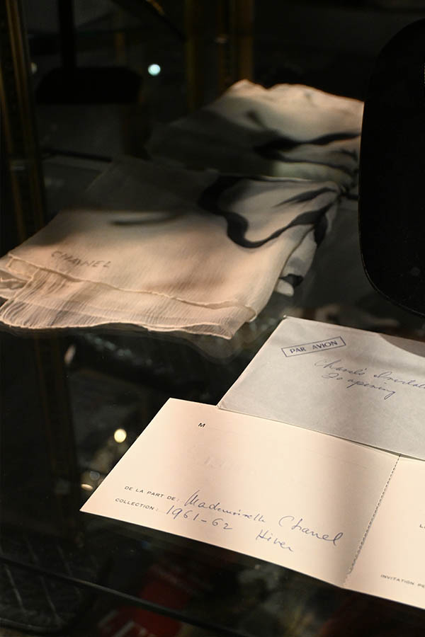 glass-display-case-shows-a-folded-sheer-Chanel-scarf,-an-envelope-marked-“PAR-AVION,”-and-an-invitation-card-from-Mademoiselle-Chanel-for-the-1961-62-Winter-Collection