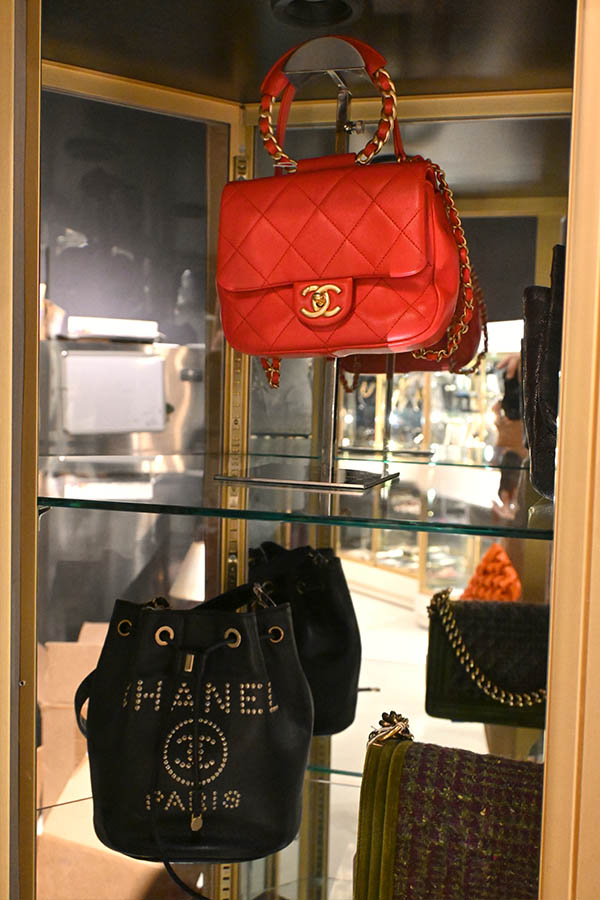 glass-display-case-shows-multiple-Chanel-designer-handbags,-including-a-red-quilted-flap-bag-and-a-black-bucket-bag-with-studded-“CHANEL-PARIS”-lettering