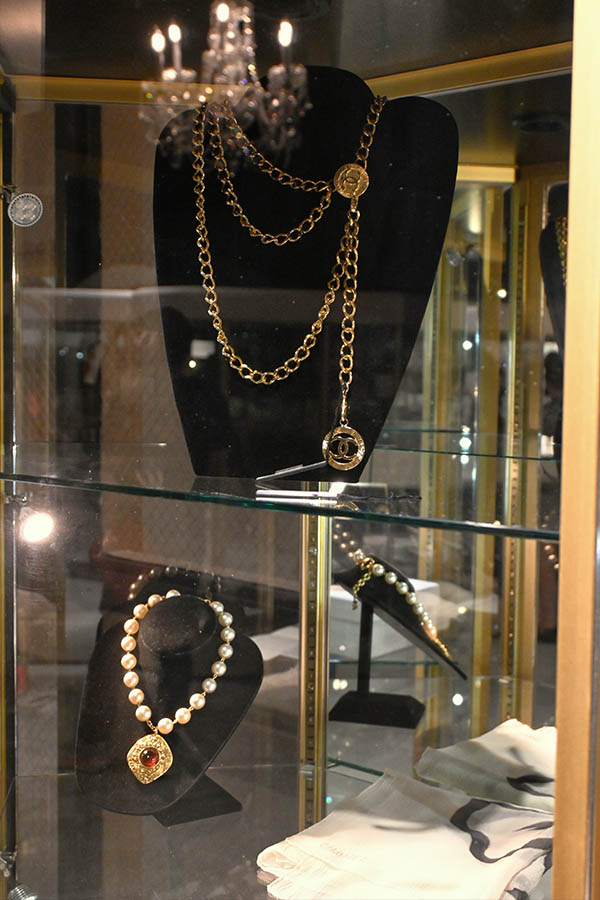 jewelry-display-case-shows-a-long-gold-chain-necklace-with-a-Chanel-logo-pendant,-a-pearl-necklace-with-a-red-pendant,-and-other-accessories-on-black-velvet-busts
