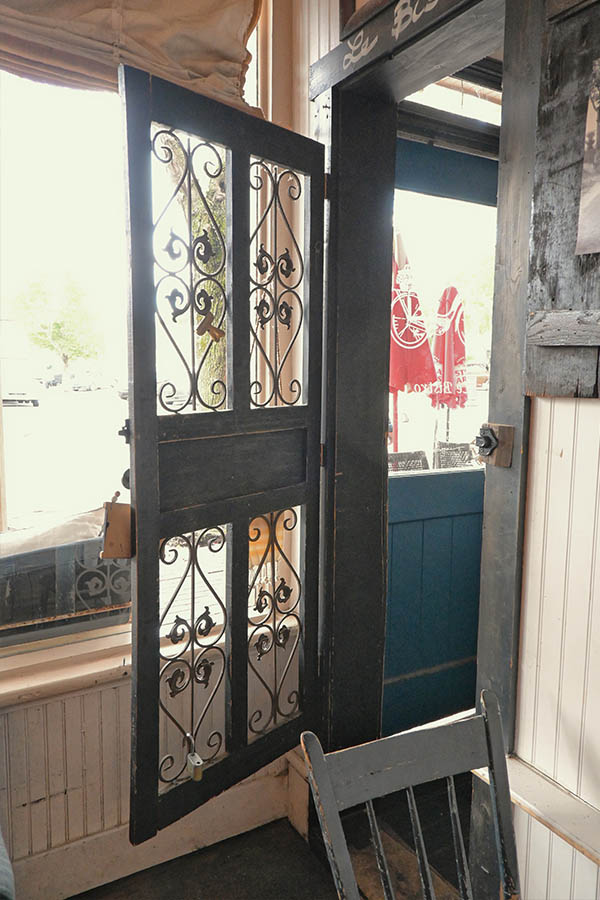 open-dark-wooden-door-with-wrought-iron-scrollwork-and-a-black-or-dark-gray-wooden-chair-in-the-foreground-in-french-restaurantt