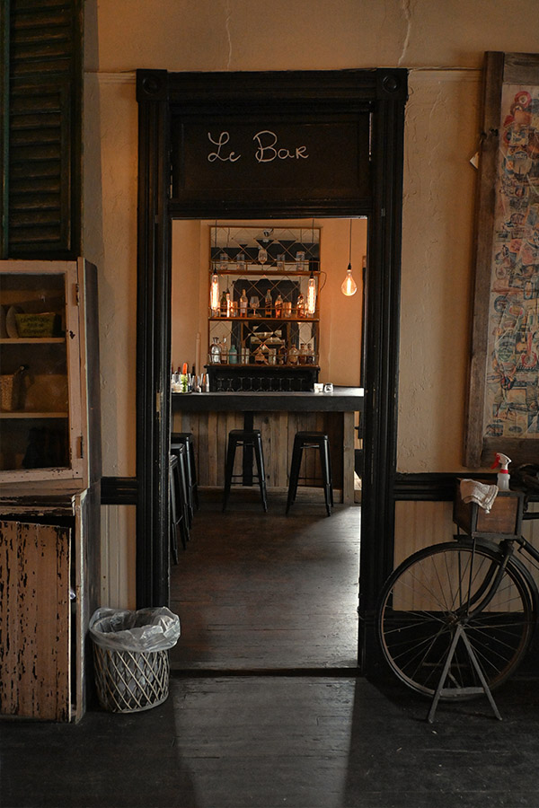 rustic-and-vintage-design-style-of-a-doorway-that-leads-into-a-bar-area-with-a-sign-above-the-door-that-says-Le-Bar-and-through-the-doorway-a-bar-counter-with-multiple-stools
