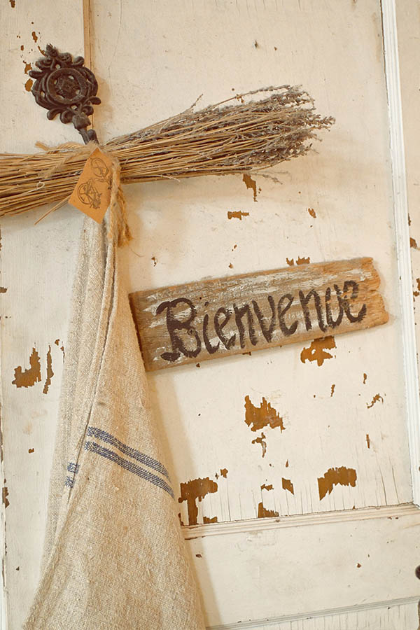 rustic-and-weathered-white-door-with-chipped-paint-and-hanging-from-it-is-a-small-wooden-plank-with-the-word-Bienvenue