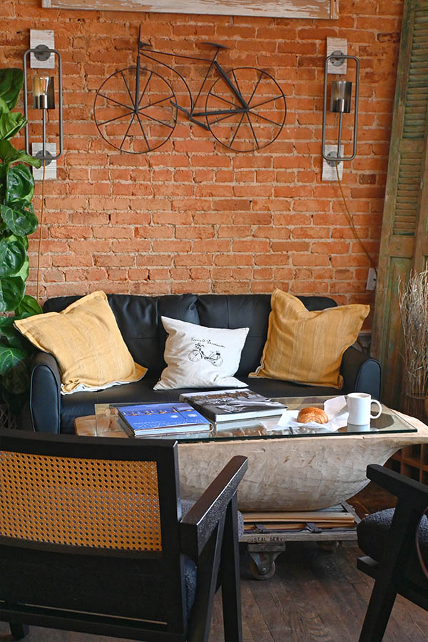 rustic-interior-space-of-Roswell-Provisions-a-textured-brick-wall-with-a-weathered-wooden-sign-and-a-wire-bicycle-sculpture-above-a-black-leather-couch-with-yellow-white-decorative-pillows