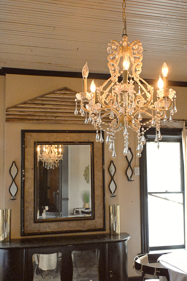rustic-interior-space-with-a-gold-or-brass-chandelier-with-crystal-pendants-hanging-from-a-white-beaded-board-ceiling-and-in-the-background-is-a-large-wooden-framed-mirror