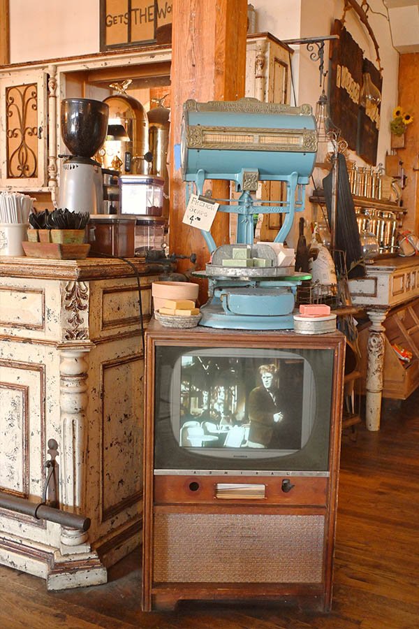 rustic-interior-space-with-a-vintage-wooden-television-set-in-the-foreground-that-is-playing-a-black-and-white-movie-or-show-and-on-top-of-the-television-is-a-large-blue-vintage-scale