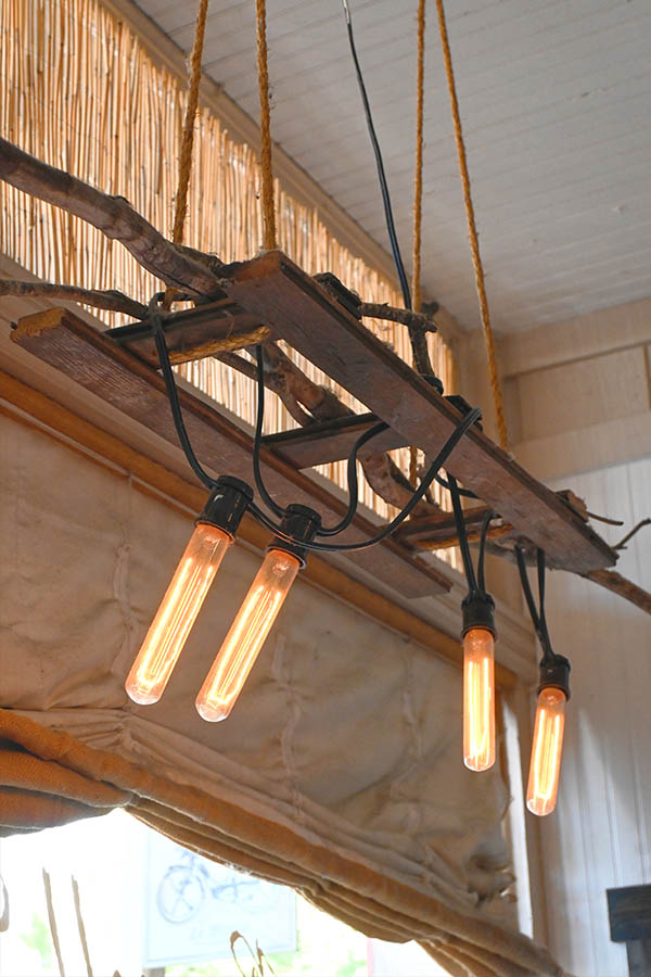 rustic-light-fixture-made-from-wooden-planks-and-branches-suspended-from-the-ceiling-by-ropes-with-multiple-exposed-filament-lightbulbs-hanging-from-black-wires-and-a-bamboo-or-reed-ceiling