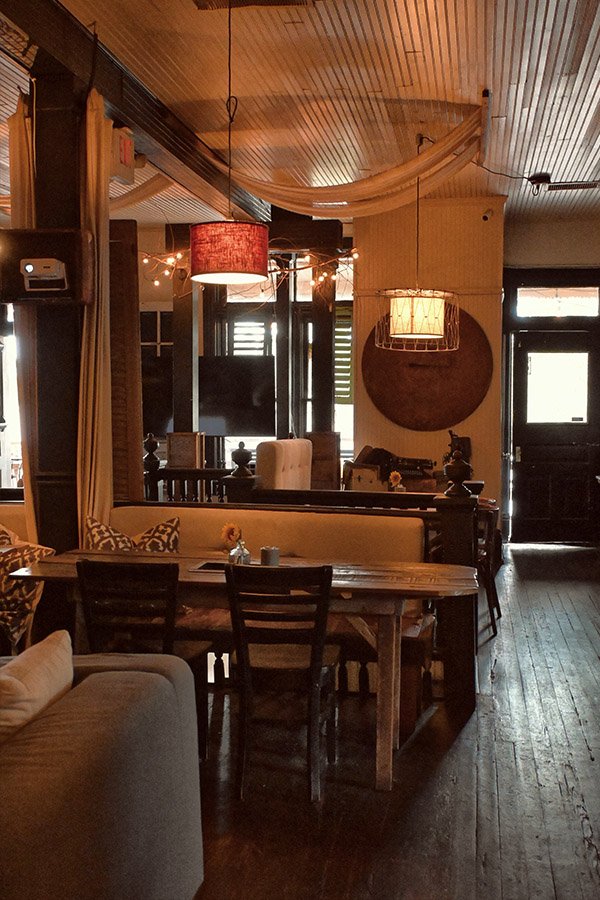 vintage-design-style-of-a-restaurant-interior-with-a-long-wooden-dining-table-and-a-built-in-bench-with-decorative-pillows-and-two-different-pendant-light-fixtures-with-fabric-shades