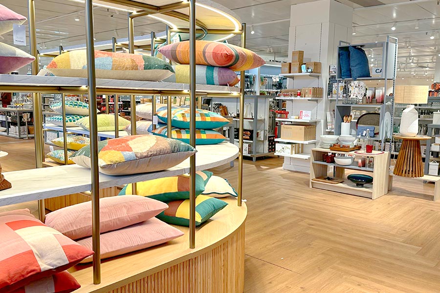 Brightly-lit-home-goods-store-featuring-a-circular-wooden-display-filled-with-colorful-patterned-pillows-and-white-marble-shelving-adjacent-to-aisles-of-modern-kitchenware-and-tabletop-decor