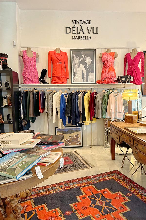Vintage-clothing-boutique-in-Marbella-featuring-mannequins-in-vibrant-pink-and-red-dresses-displayed-above-racks-of-colorful-jackets-with-stacks-of-art-books