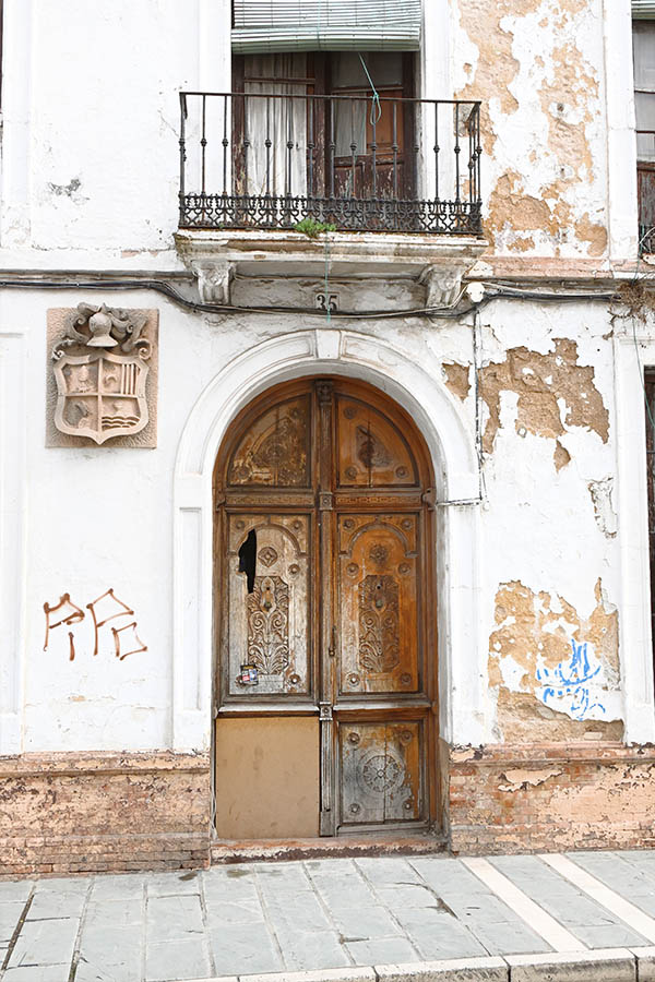 arched-wooden-door-with-ornate-carvings-set-into-a-weathered-white-facade-featuring-a-stone-coat-of-arms-and-a-wrought-iron-balcony-above
