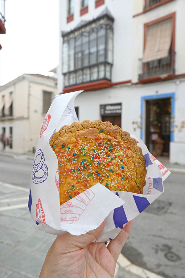 hand-holding-a-round-golden-brown-cookie-topped-with-colorful-rainbow-sprinkles-wrapped-in-patterned-white-paper