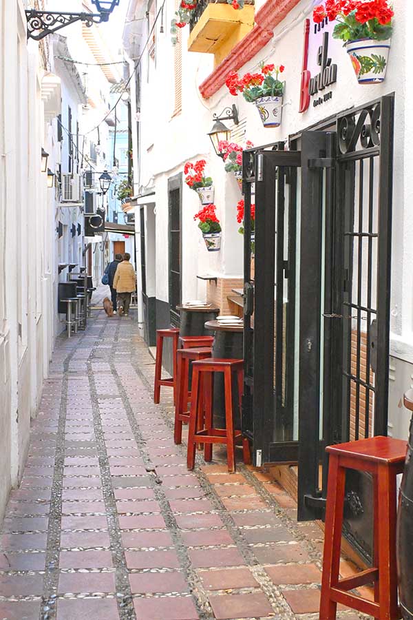 narrow-whitewashed-alleyway-features-red-potted-flowers-on-balconies-and-walls-with-several-tall-red-wooden-stools-marbella-spain