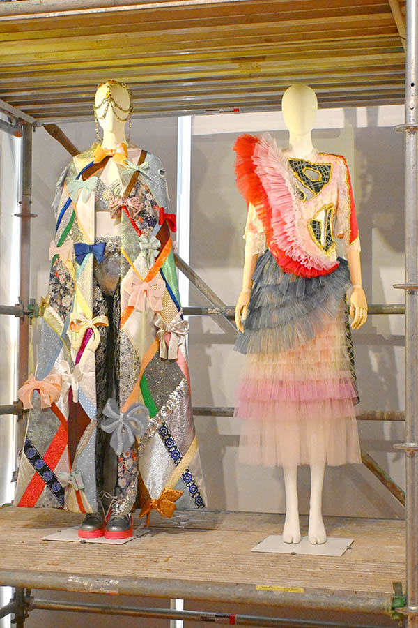 patchwork-couture-cape-with-multi-colored-bows-beside-pink-tulle-dress-on-scaffolding