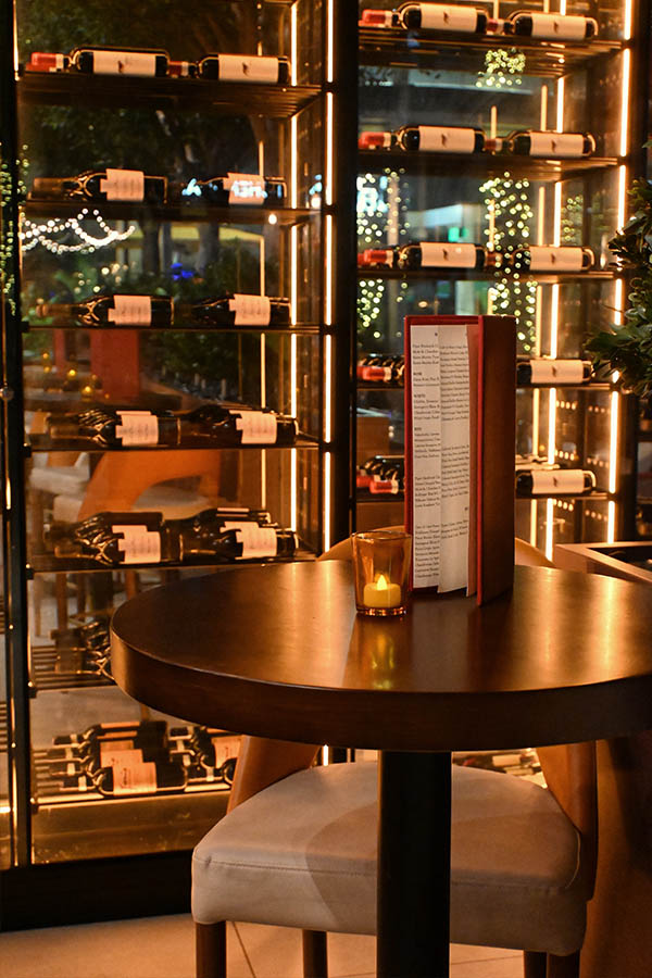 small-round-wooden-table-with-a-flickering-candle-and-a-red-menu-stands-in-front-of-tall-glass-wine-refrigerators-filled-with-horizontal-bottles-illuminated-by-strips-of-warm-light