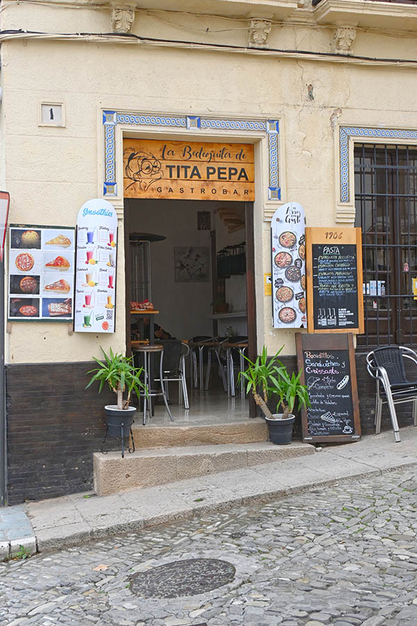 the-entrance-to-a-gastrobar-named-la-bodeguita-de-tita-pepa-featuring-an-open-doorway-flanked-by-potted-plants-and-menu-boards-displaying-pizzas-smoothies