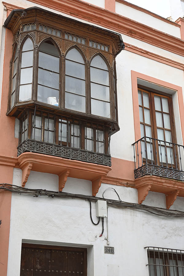 white-exterior-wall-of-a-building-featuring-an-ornate-wooden-bay-window-with-pointed-arches-and-an-intricate-wrought-iron-railing