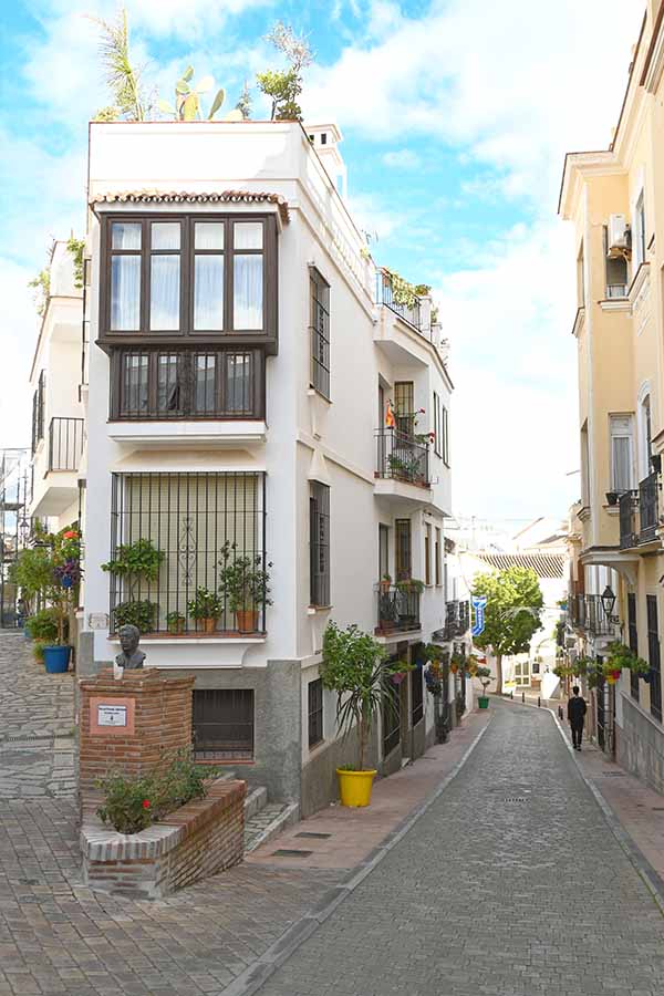 Narrow-white-building-on-a-street-corner-with-a-brown-wooden-balcony-overlooking-a-cobblestone-path