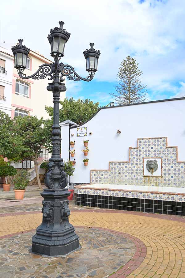 Ornate-black-lamppost-stands-before-a-white-wall-with-a-tiled-lion-fountain-and-potted-plants-under-a-bright-sky