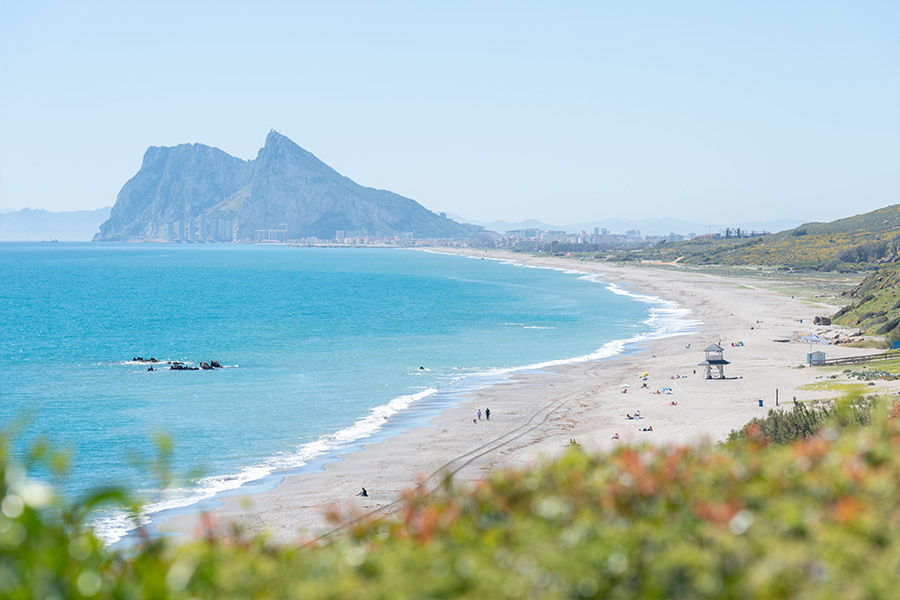 The-Rock-of-Gibraltar-anchors-the-horizon-line-of-a-long-curving-beach-under-a-clear-sky