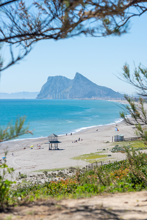 The-Rock-of-Gibraltar-stands-at-the-end-of-a-sunny-sandy-beach-with-a-white-lifeguard-tower